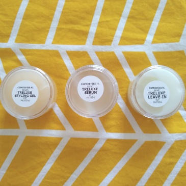 Three small transparent tubs on a yellow background with white diagonal lines. From left to right: the TréLuxe Styling Gel, TréLuxe Serum and TréLuxe Leave-In
