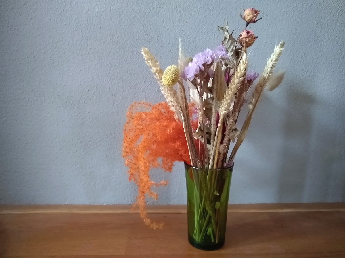 A photo of a dried flower bouquet in beige, orange, pinks and purples, in a green glass, standing on a wooden floor in front of a grey wall.