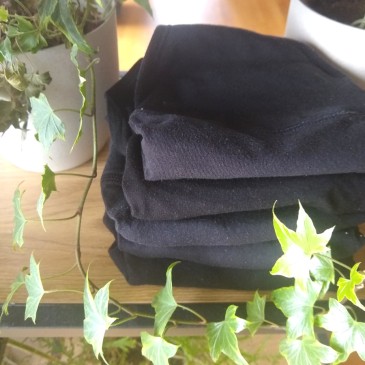 A pile of 5 folded black period panties, on a wooden shelf with plants surrounding them.