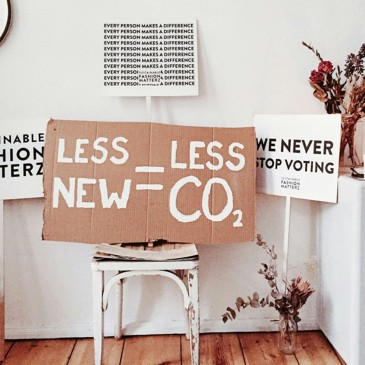 A photo of a white room with white furniture. On a chair there is a cardboard sign that reads "Less New = Less CO2". A sign on the left reads "Sustainable fashion matterz", a sign on the right reads "We never stop voting", and a sign above reads "Every person makes a difference" multiple times.